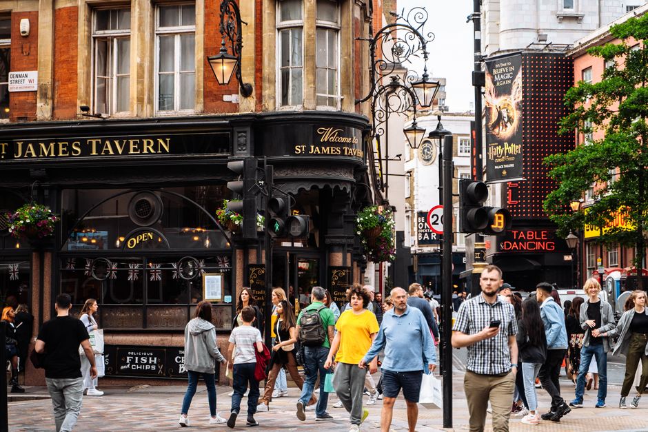 The image depicts a busy urban street scene on a cloudy day, featuring a mix of historic and modern architecture with ornate facades and large windows, typical of central London. In the foreground, se