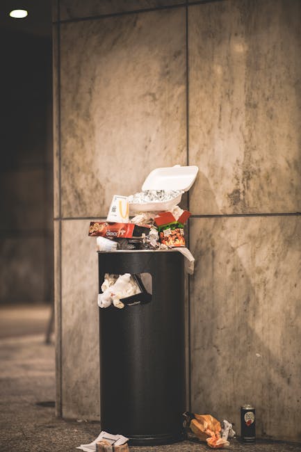 A narrow, urban alleyway filled with overflowing waste and discarded rubbish bags, with a large black sack in the foreground pulled by a trolley on the uneven, dirty pavement. Behind the trolley, pile