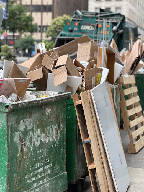 A collection of overflowing rubbish bins and scattered waste bags on a paved sidewalk adjacent to a parking area. The waste containers include a large grey mixed paper and card bin with an open lid, r